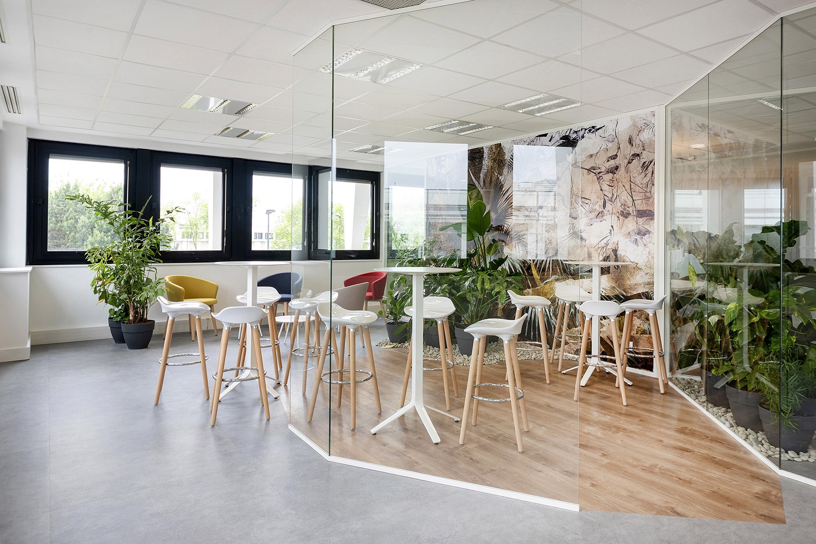 Espace de pause moderne avec tables hautes, tabourets blancs, cloisons en verre et plantes d'intérieur en abondance, avec de grandes fenêtres et de la lumière naturelle.