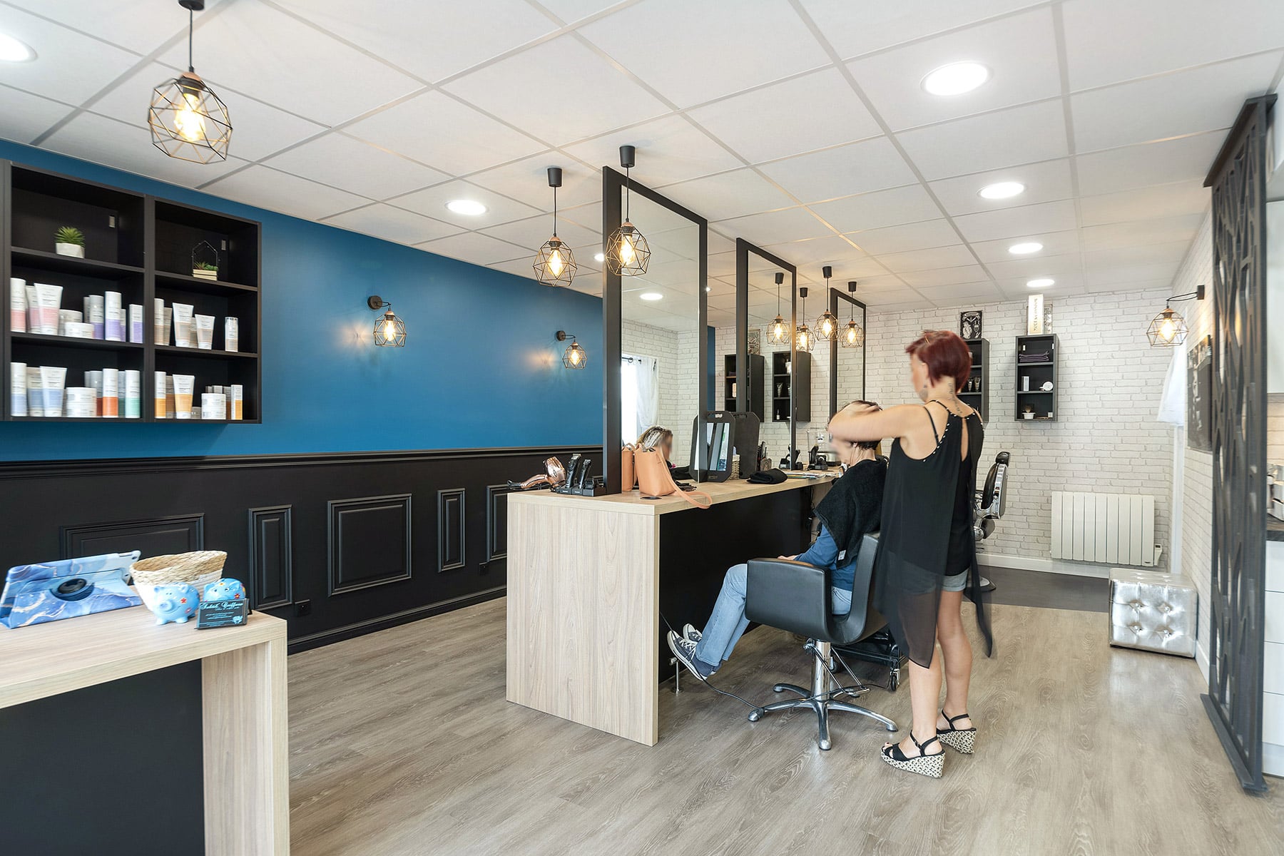 Un coiffeur lave les cheveux d'une cliente dans un salon moderne aux murs bleus et blancs, au sol en bois et aux étagères présentant des produits capillaires.