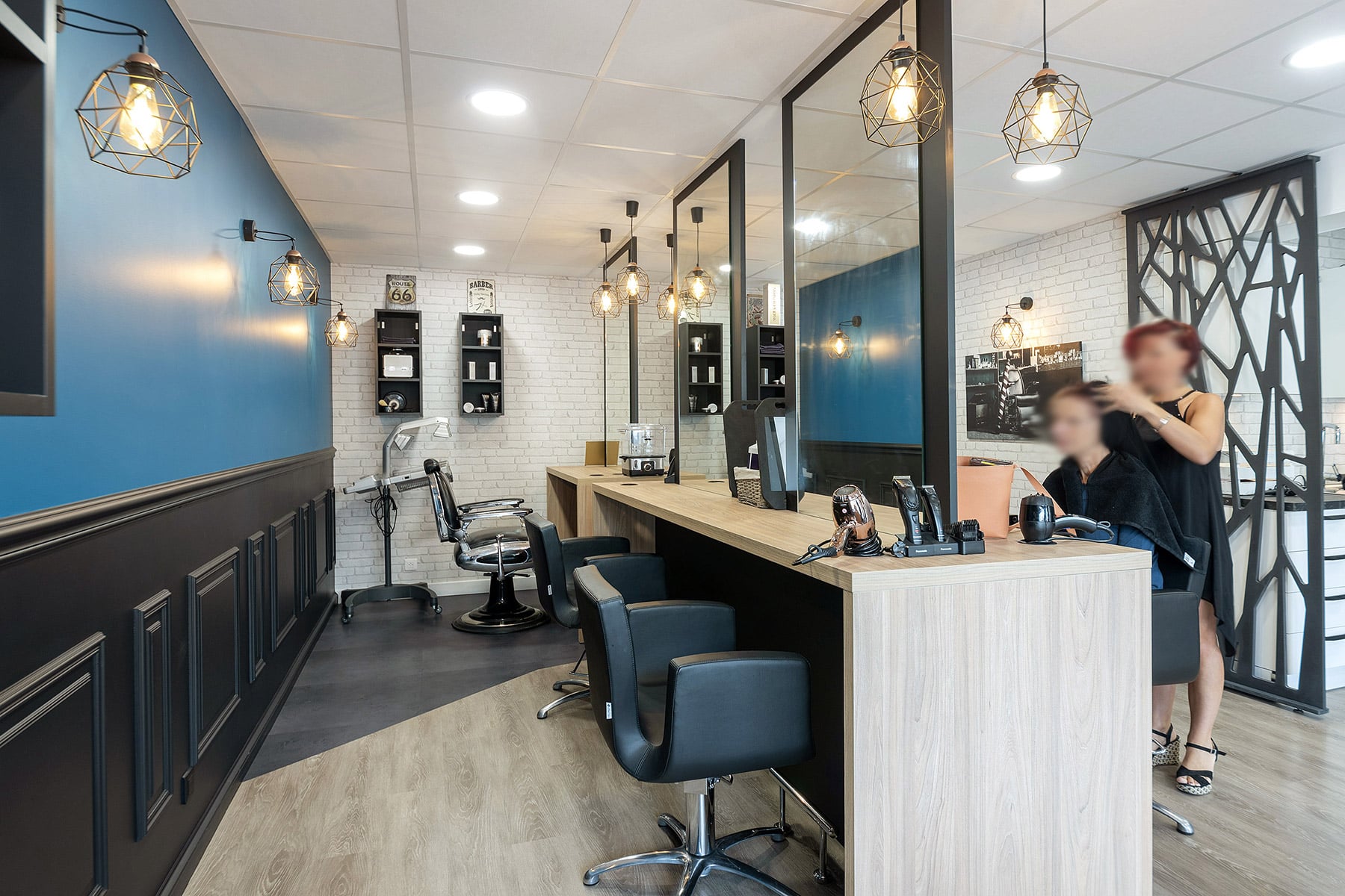 Intérieur d'un salon de coiffure moderne avec des chaises noires, de grands miroirs et des lampes suspendues ; un coiffeur travaille sur les cheveux d'une cliente.
