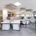 Bureau moderne à aire ouverte avec des bureaux blancs, des chaises ergonomiques, des panneaux de séparation, des plantes en pot et une lumière vive. Aucune personne n'est présente.