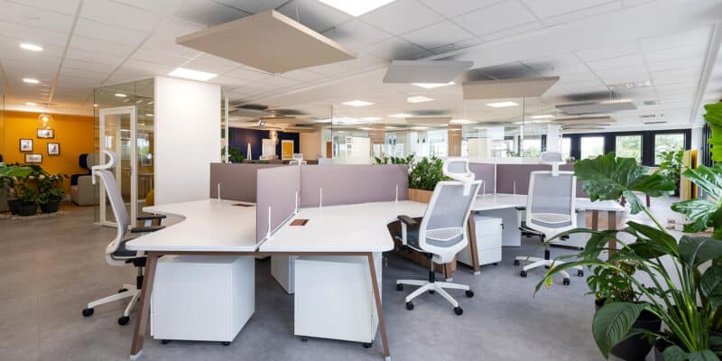 Bureau moderne à aire ouverte avec des bureaux blancs, des chaises ergonomiques, des panneaux de séparation, des plantes en pot et une lumière vive. Aucune personne n'est présente.