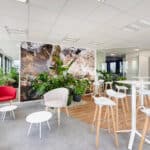 Salon de bureau moderne avec des chaises et des tables blanches, des fauteuils colorés, des plantes d'intérieur et de grandes fenêtres laissant entrer la lumière naturelle.