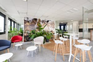 Salon de bureau moderne avec des chaises et des tables blanches, des fauteuils colorés, des plantes d'intérieur et de grandes fenêtres laissant entrer la lumière naturelle.
