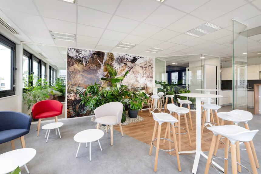 Salon de bureau moderne avec des chaises et des tables blanches, des fauteuils colorés, des plantes d'intérieur et de grandes fenêtres laissant entrer la lumière naturelle.