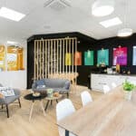 Salle de pause d'un bureau moderne avec une table en bois, des chaises blanches, des sièges de salon, une kitchenette, des affiches colorées sur un mur noir et un réfrigérateur orange.