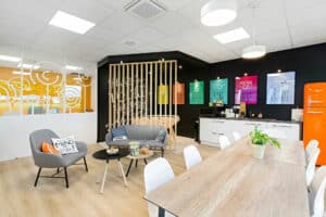 Salle de pause d'un bureau moderne avec une table en bois, des chaises blanches, des sièges de salon, une kitchenette, des affiches colorées sur un mur noir et un réfrigérateur orange.