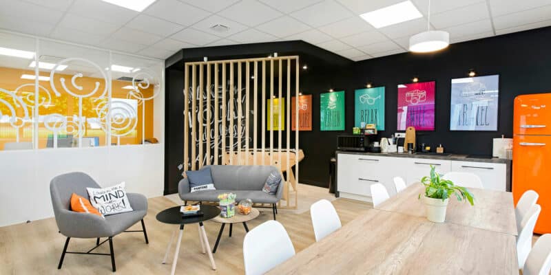 Salle de pause d'un bureau moderne avec une table en bois, des chaises blanches, des sièges de salon, une kitchenette, des affiches colorées sur un mur noir et un réfrigérateur orange.