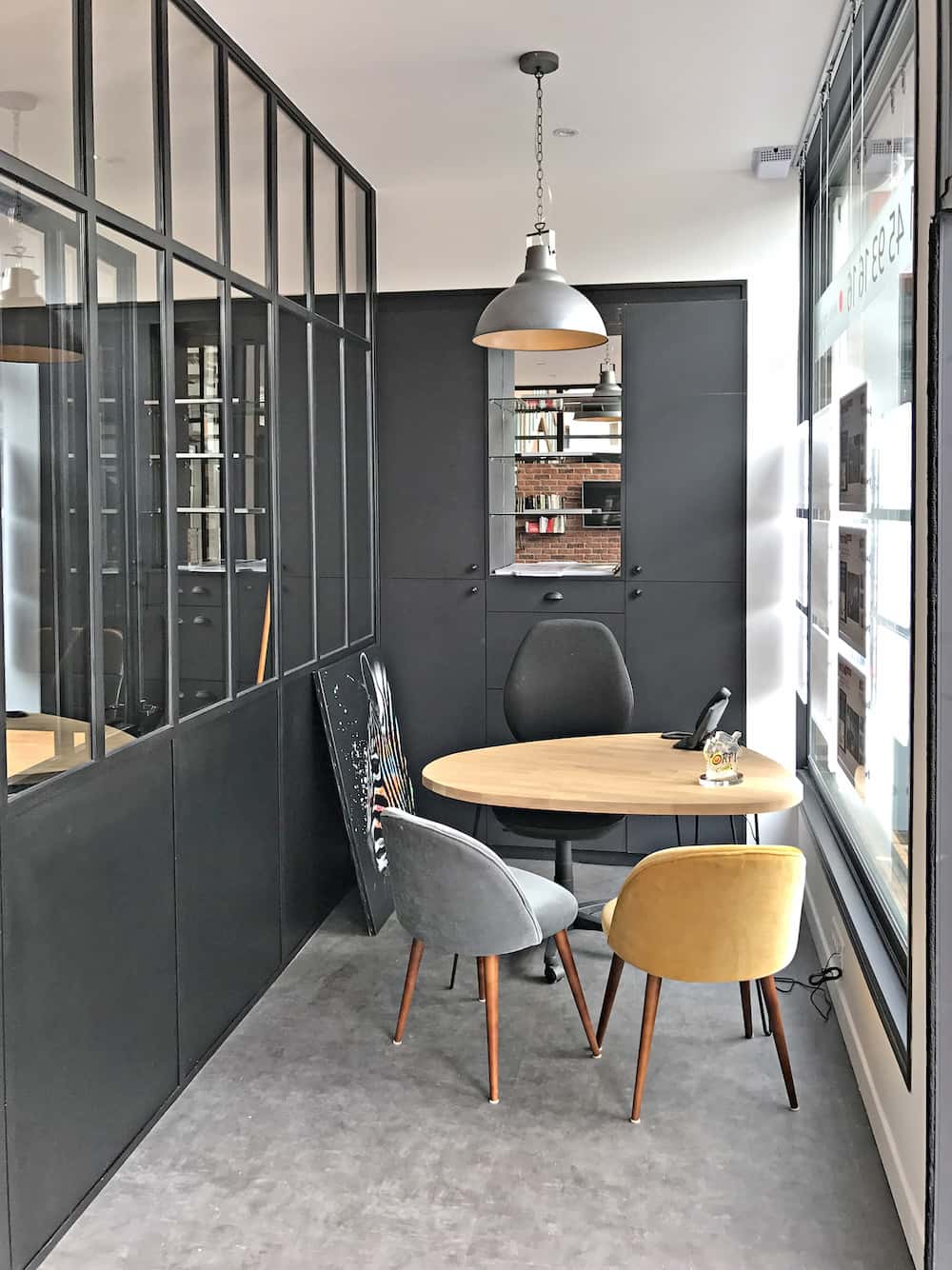 Un petit bureau moderne avec des cloisons en verre, une table ronde et trois chaises de couleurs différentes présente un agencement de bureau élégant. Une lampe suspendue et des armoires murales complètent l'ensemble, avec un téléphone et un verre sur la table.