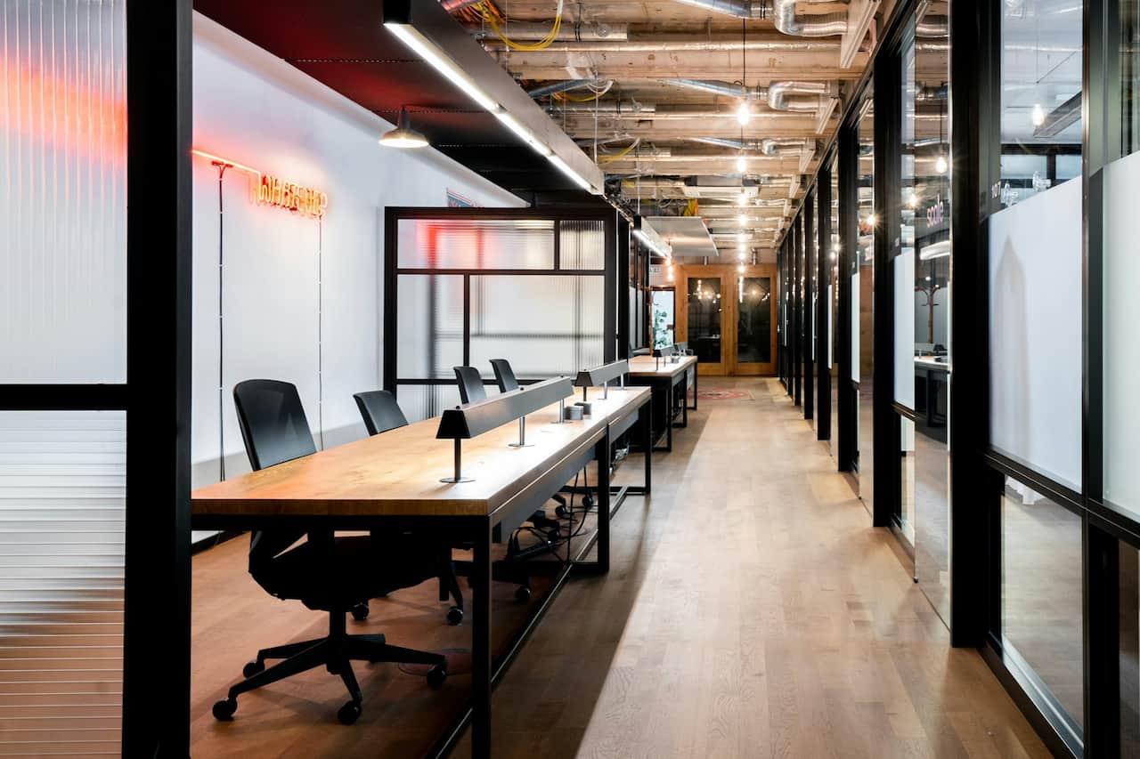 Bureau moderne à aire ouverte avec de longs bureaux partagés, des chaises noires, des lampes de bureau, des murs en verre et des tuyaux de plafond apparents, éclairés par un éclairage naturel et artificiel.
