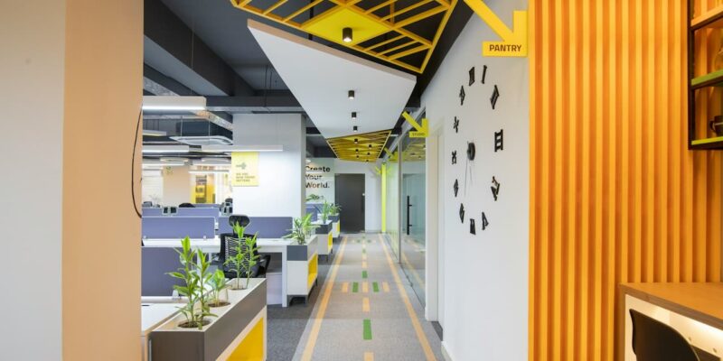 Couloir de bureau moderne avec un plafond géométrique, des plantes d'intérieur, un panneau directionnel de garde-manger et des cabines visibles sur la gauche.