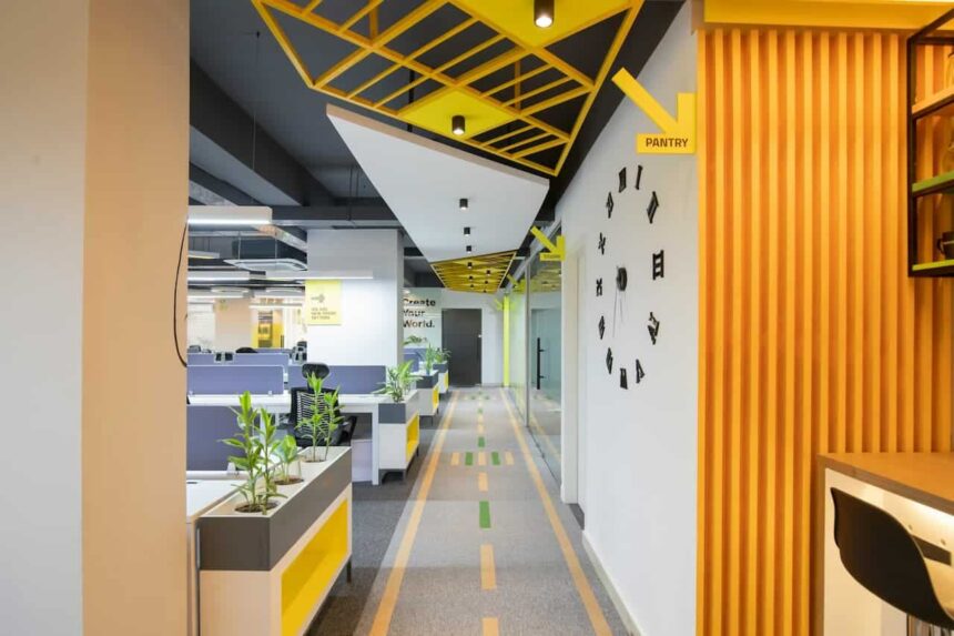 Couloir de bureau moderne avec un plafond géométrique, des plantes d'intérieur, un panneau directionnel de garde-manger et des cabines visibles sur la gauche.