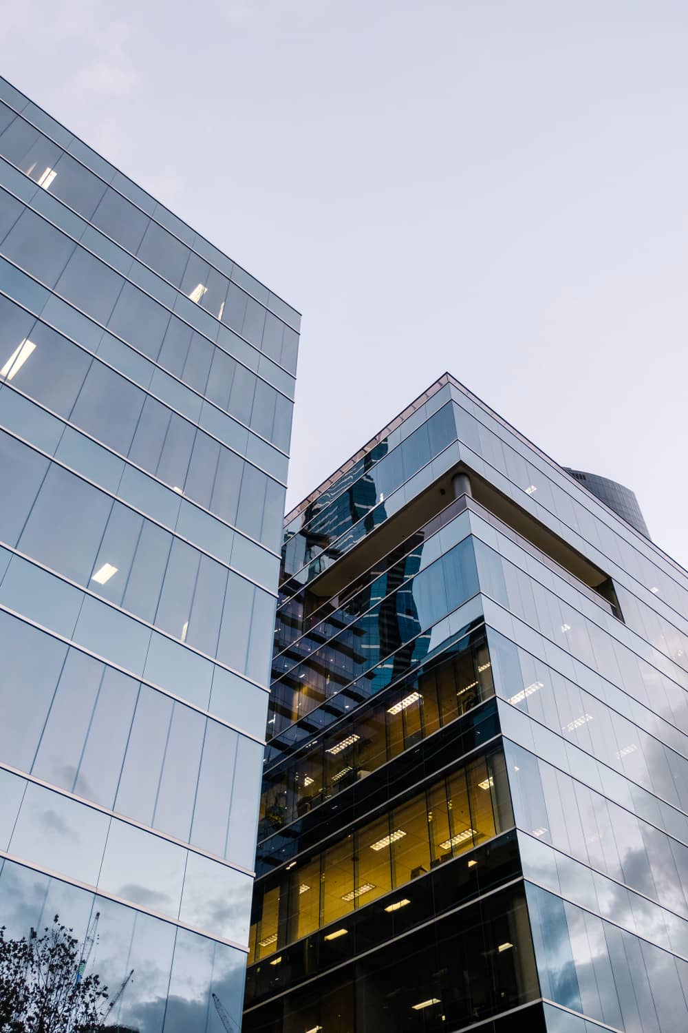 Des immeubles de bureaux modernes en verre reflétant le ciel et les structures voisines, avec des lumières intérieures visibles à travers certaines fenêtres, mettent en valeur un agencement de bureau élégant, photographié depuis un angle bas.