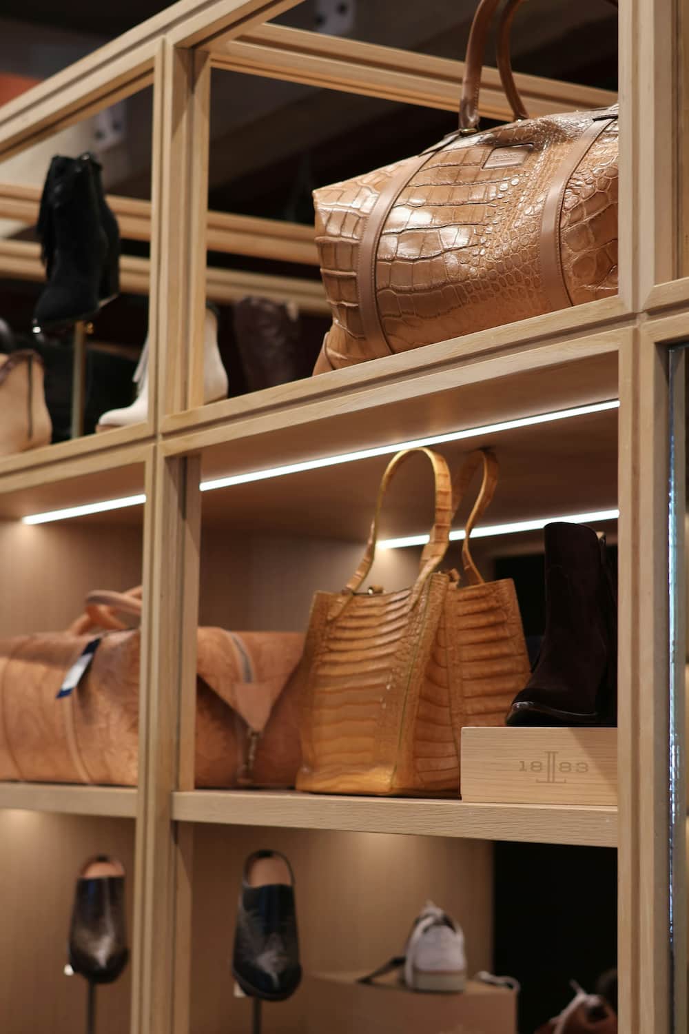 Des étagères présentent divers sacs à main et bottes en cuir, dont un sac de sport et un fourre-tout à motif crocodile, dans une boutique bien éclairée aux influences élégantes d'agencement de bureau.