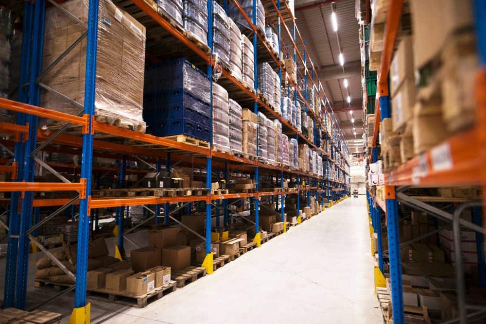 Intérieur d'un entrepôt avec de hautes étagères bleues et orange remplies de palettes et de boîtes, avec une allée spacieuse - une configuration industrielle qui contraste avec l'agencement de bureau organisé que l'on trouve dans les environnements de bureau.