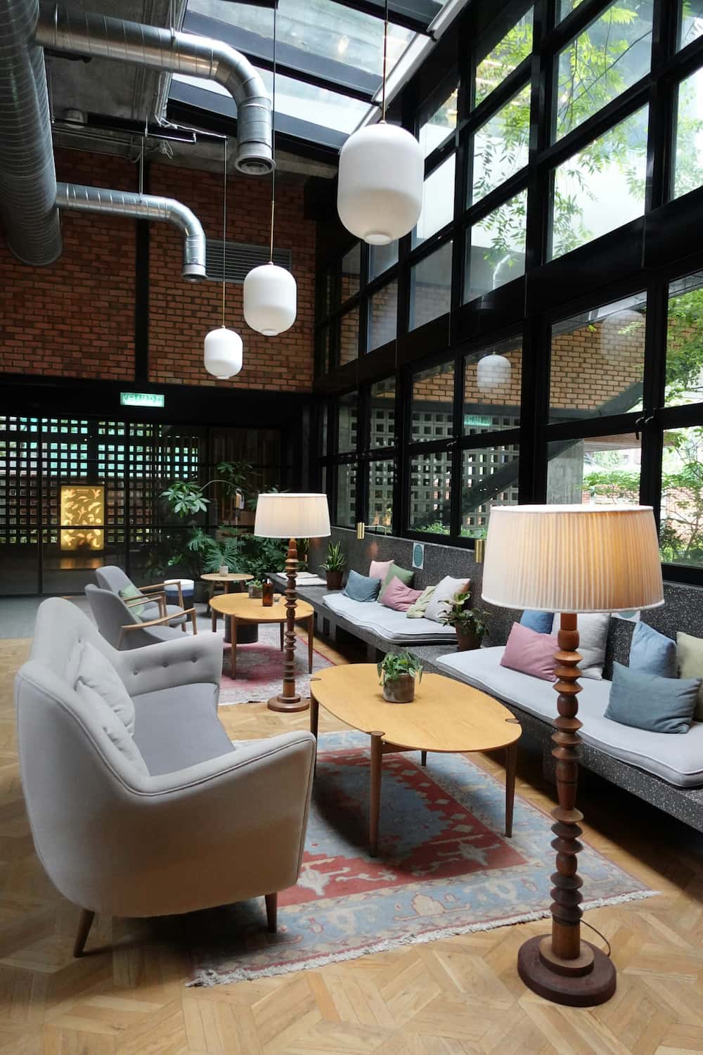 Salon moderne avec de grandes fenêtres, des canapés, des fauteuils, des tables en bois, des lampadaires, des plantes d'intérieur et des lampes suspendues sous un plafond industriel - idéal pour un agencement de bureau élégant.
