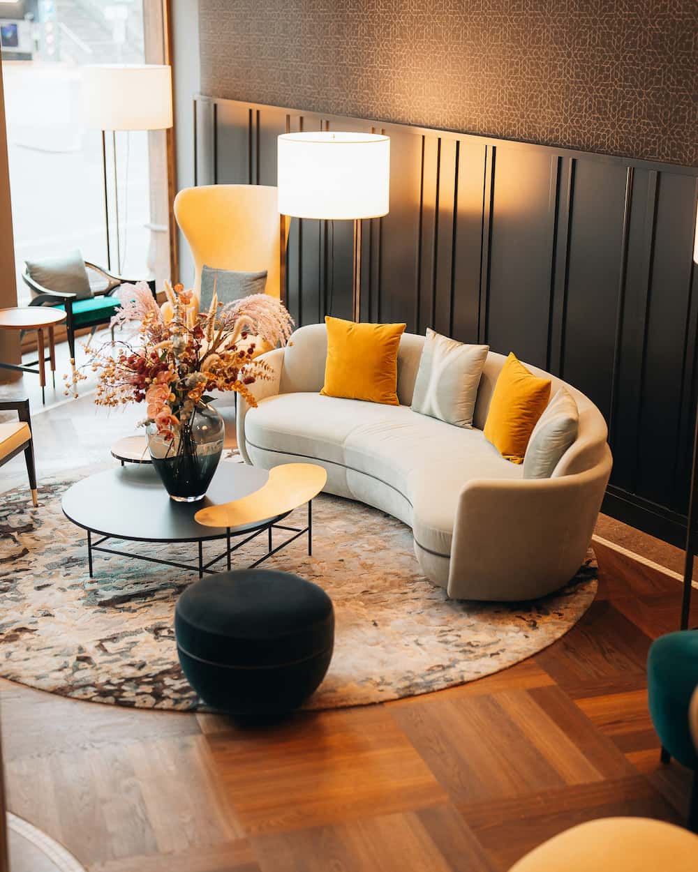 Un salon moderne avec un canapé beige incurvé, des coussins jaunes et gris, une table basse ronde avec un arrangement floral, deux lampadaires et un tapis à motifs sur un sol en bois - une inspiration parfaite pour un agencement de bureau élégant et accueillant.