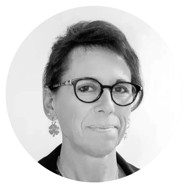 Une personne aux cheveux courts et noirs, portant des lunettes et des boucles d'oreilles en forme de trèfle, regarde l'appareil photo avec une expression neutre. L'image est en noir et blanc.