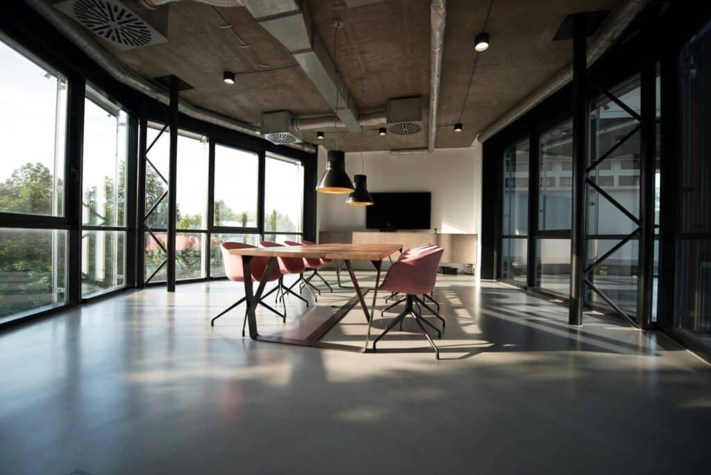 Salle de conférence moderne avec une table en bois, des chaises roses, de grandes fenêtres, des plafonniers, une télévision et des conduits apparents au plafond. La lumière du soleil pénètre à l'intérieur - un exemple inspirant d'agencement bureaux Île-de-France par un contractant général rénovation compétent.