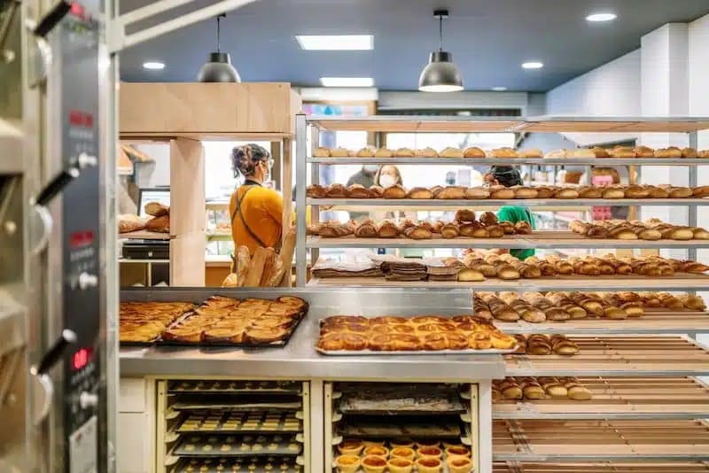 L'intérieur de la boulangerie présente un agencement expert en boulangerie avec des étagères de pains et de pâtisseries assortis, du personnel travaillant à l'arrière-plan et des plateaux de produits de boulangerie au premier plan.