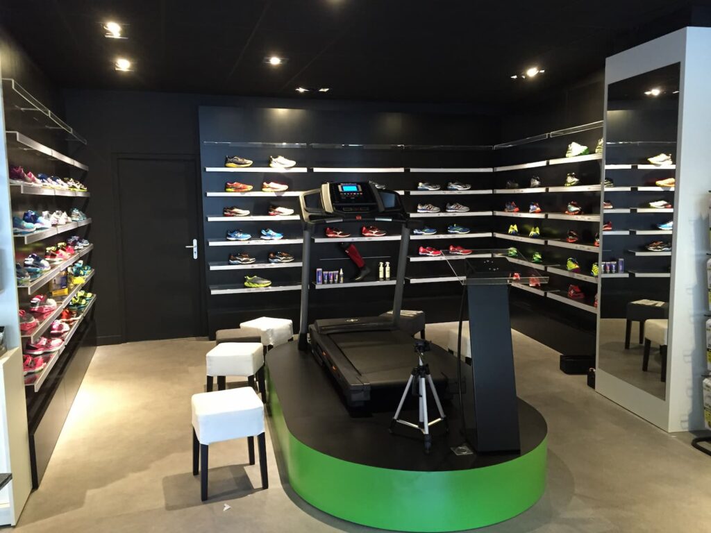 Un magasin de chaussures moderne présente des étalages muraux de chaussures de sport, un tapis roulant sur une plate-forme surélevée, des tabourets et un trépied dans un intérieur minimaliste et bien éclairé.