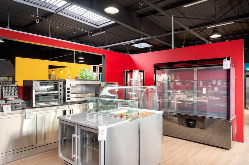 Exposition d'une cuisine commerciale comprenant des fours en acier inoxydable, des vitrines, un comptoir réfrigéré et des étagères sur des murs rouges et jaunes.