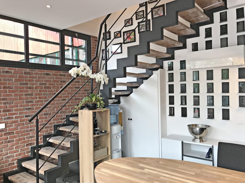 Intérieur de loft moderne avec un escalier en bois et métal noir, un mur de briques apparentes et une galerie de photos sur le mur blanc. Une table et une étagère en bois se trouvent au premier plan.
