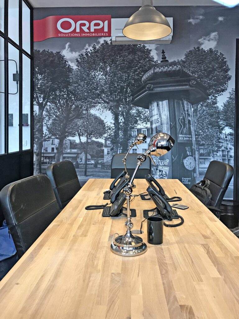Une salle de conférence moderne avec des chaises noires, des téléphones de bureau, des tasses et une grande peinture murale d'une scène de rue parisienne ; un panneau "ORPI Solutions Immobilières" sur le mur.