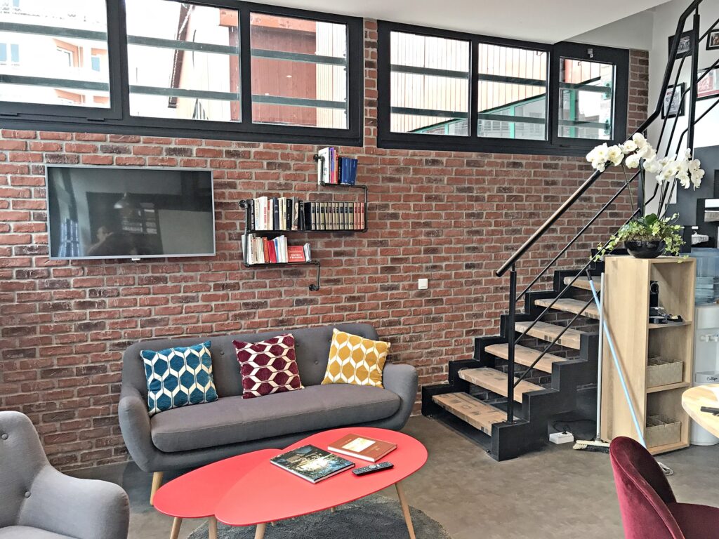 Salon moderne avec mur en briques apparentes, canapé avec coussins à motifs, télévision murale, étagères, table basse rouge et escalier avec rampe en métal.