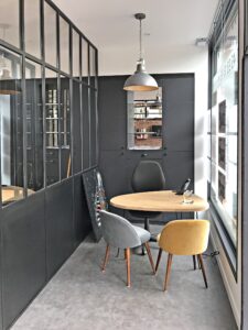Un bureau moderne avec une table ronde, trois chaises dépareillées, une lampe suspendue, des armoires sombres, une cloison en verre et une fenêtre laissant entrer la lumière naturelle.