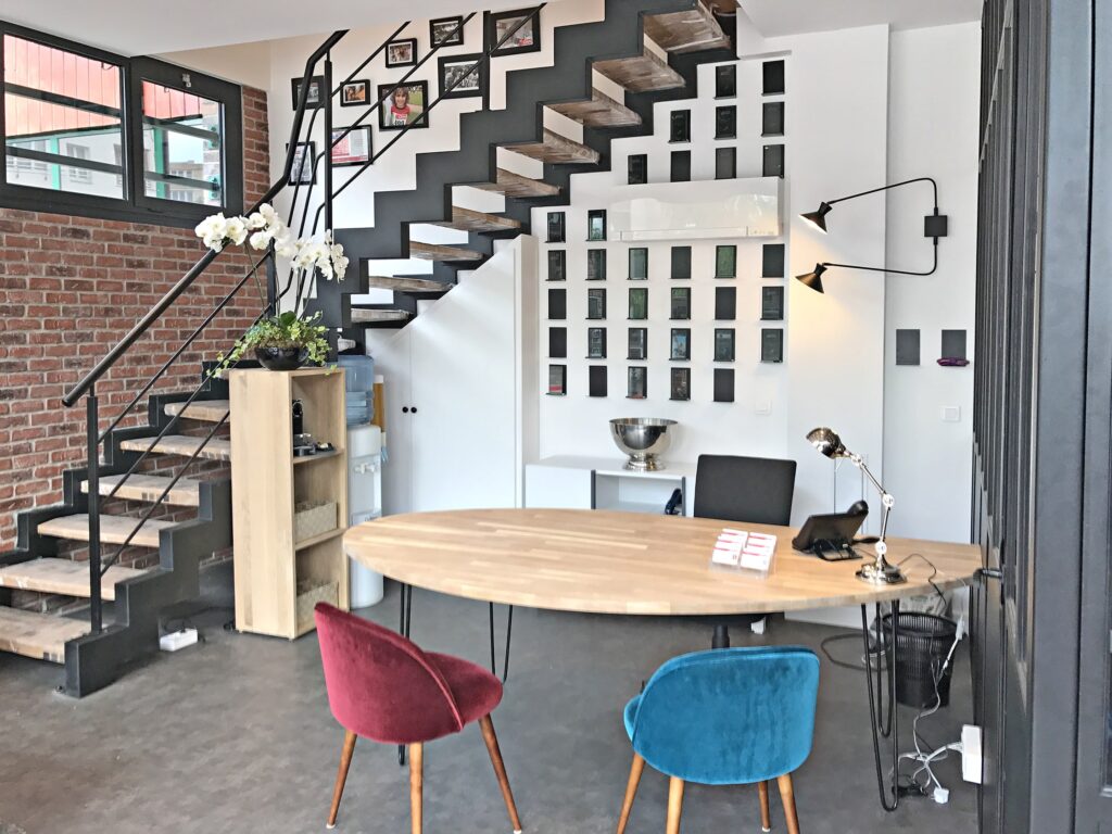 Un intérieur de bureau moderne avec un bureau en bois, des chaises colorées, un escalier en métal et des images encadrées au mur.