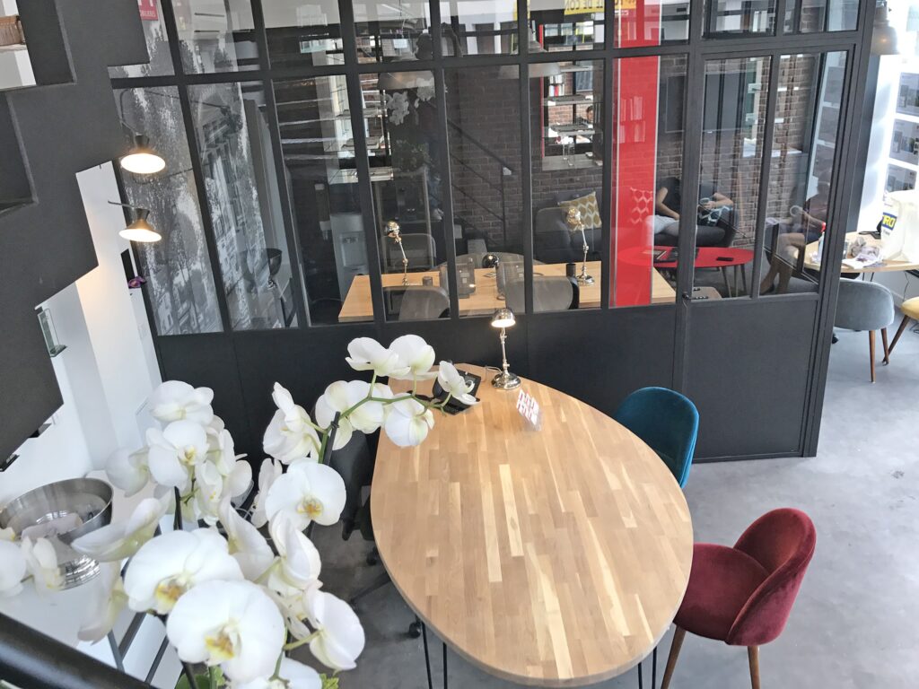 Bureau moderne avec des murs en verre, une longue table en bois, des chaises colorées et des orchidées blanches au premier plan. Plusieurs zones de travail sont visibles derrière les cloisons en verre.