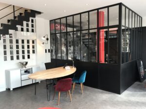 Bureau moderne avec une cloison en verre, un bureau ovale en bois, des chaises colorées, un buffet blanc et un escalier en métal noir avec des cadres photo accrochés au mur.