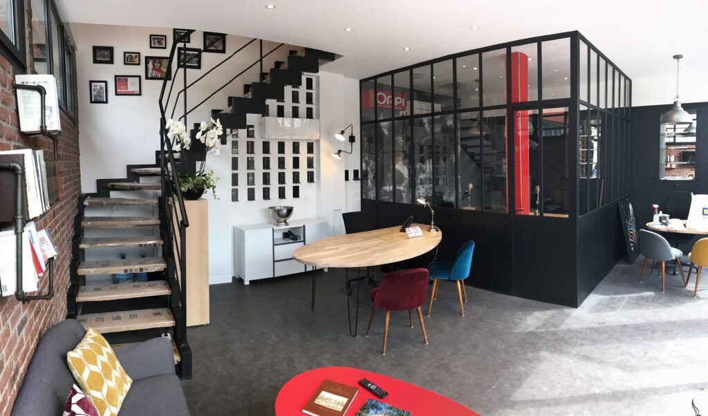 Espace de bureau moderne avec un agencement ouvert, une salle de réunion vitrée, des chaises colorées autour d'une table en bois, un escalier et des accents décoratifs sur les murs.