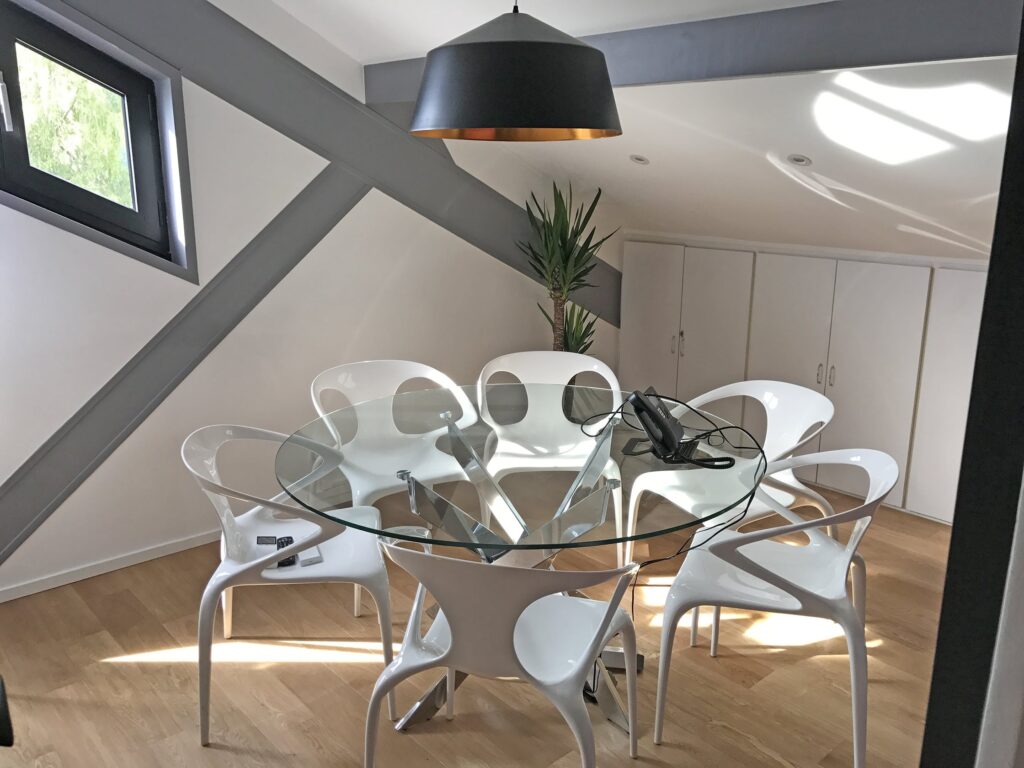 Salle de réunion moderne avec une table ronde en verre, six chaises blanches, une grande lampe suspendue noire et des armoires sous un plafond incliné. Une plante est posée dans un coin.