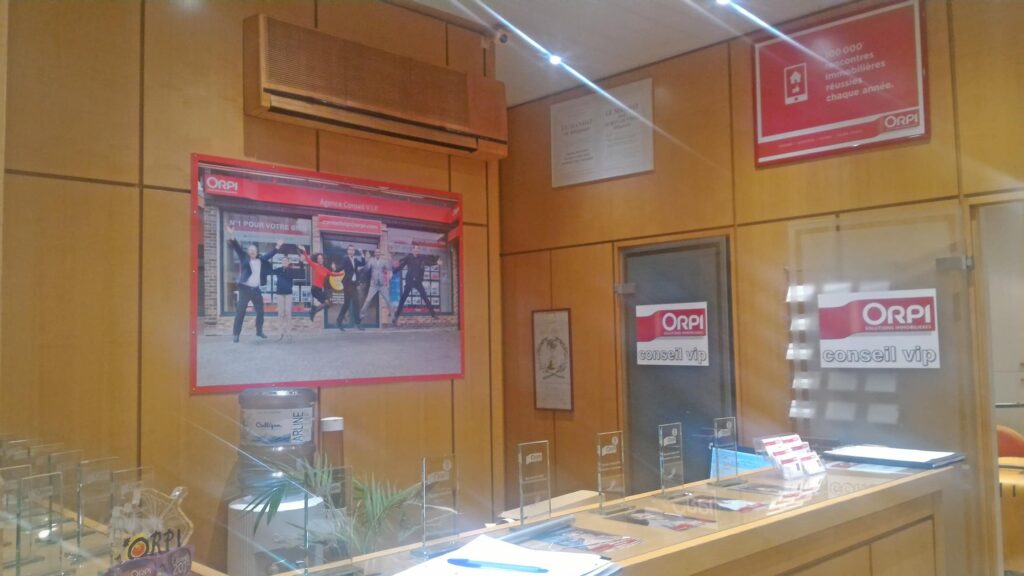 Réception d'un bureau avec des murs en bois, la marque ORPI, une grande photo sur le mur, des brochures sur le comptoir et des cloisons en verre portant la mention "conseil vip".