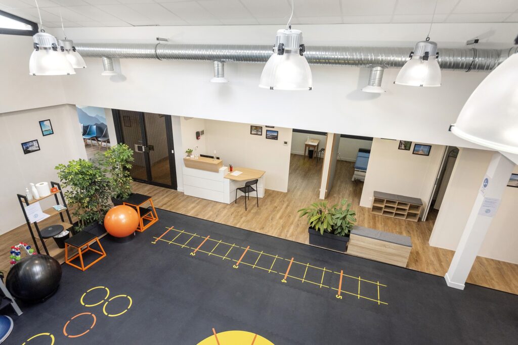 Une salle de sport ou un studio de fitness moderne avec des ballons d'exercice, des plantes d'intérieur, un bureau d'accueil et un sol noir marqué de lignes et de cercles d'entraînement jaunes et orange.