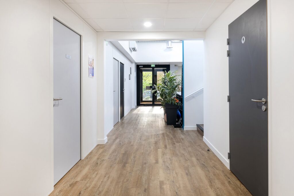 Un couloir propre et moderne avec un sol en bois, des murs blancs, plusieurs portes fermées, des plantes en pot et une porte de sortie en verre au bout qui laisse entrer la lumière naturelle.