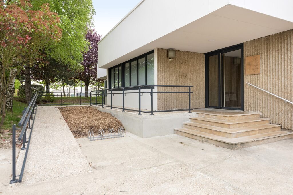 Une entrée de bâtiment moderne avec une rampe, des marches, des mains courantes en métal et un support à vélos, entourée d'arbres et d'une petite zone paillée.