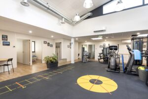 Salle de sport moderne avec des machines d'exercice, des stations d'entraînement, une échelle d'agilité jaune au sol, des plantes en pot et des photos encadrées sur les murs blancs sous des plafonniers lumineux.