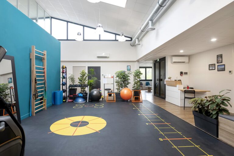 Salle de sport moderne équipée de matériel d'exercice, de ballons d'équilibre, d'une échelle murale, de marquages au sol pour les exercices d'agilité, de miroirs, de plantes et d'un bureau d'accueil dans un espace ouvert et lumineux.