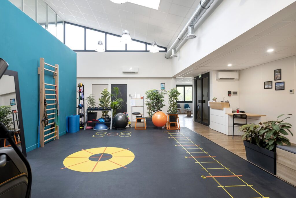 Une salle de sport moderne avec des appareils d'exercice, des ballons de médecine colorés, des barres murales, des plantes, de grandes fenêtres et un bureau d'accueil à l'arrière-plan.