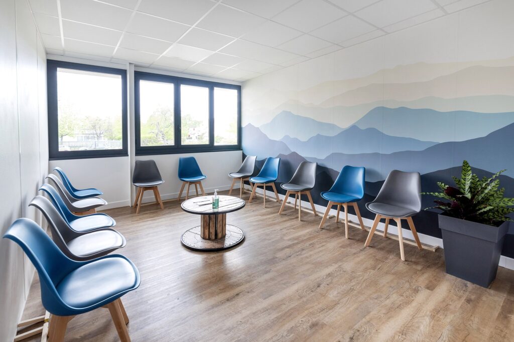 Une salle d'attente moderne avec des chaises bleues et grises disposées le long des murs, une table ronde au centre, une plante en pot et une peinture murale de montagnes abstraites.
