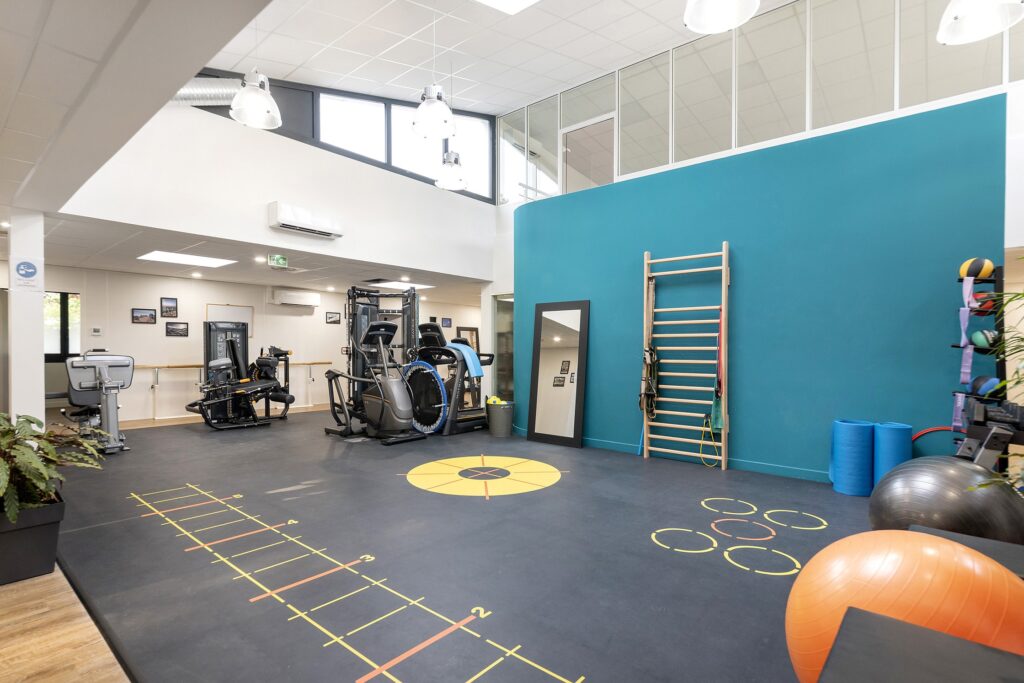 Salle de sport spacieuse équipée d'appareils d'exercice, d'un mur d'escalade, de marquages au sol, de tapis de yoga, de ballons de médecine et de grandes fenêtres laissant entrer la lumière naturelle.