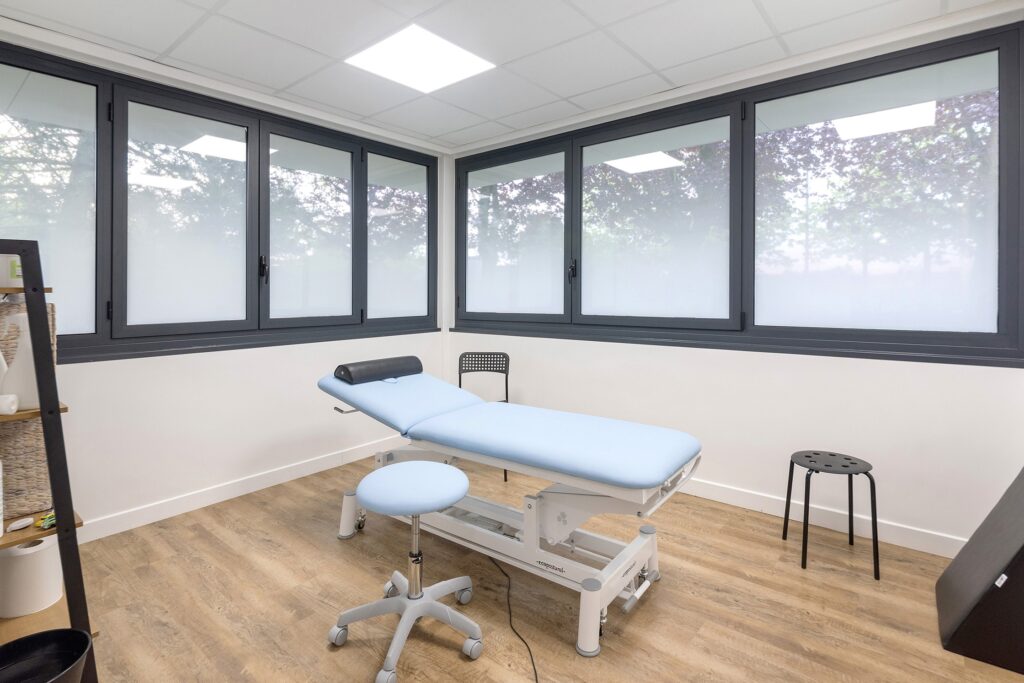 Une salle d'examen médical avec une table d'examen bleu clair, un tabouret bleu, un tabouret noir et de grandes fenêtres en verre dépoli.