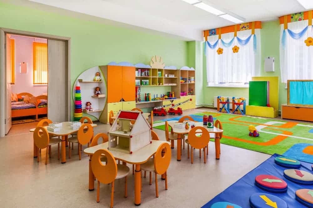 Une salle de classe préscolaire bien éclairée avec de petites tables et des chaises, des étagères colorées avec des jouets, une maison de jeu et une aire de jeu avec un tapis. Une salle de sieste est visible par une porte ouverte.