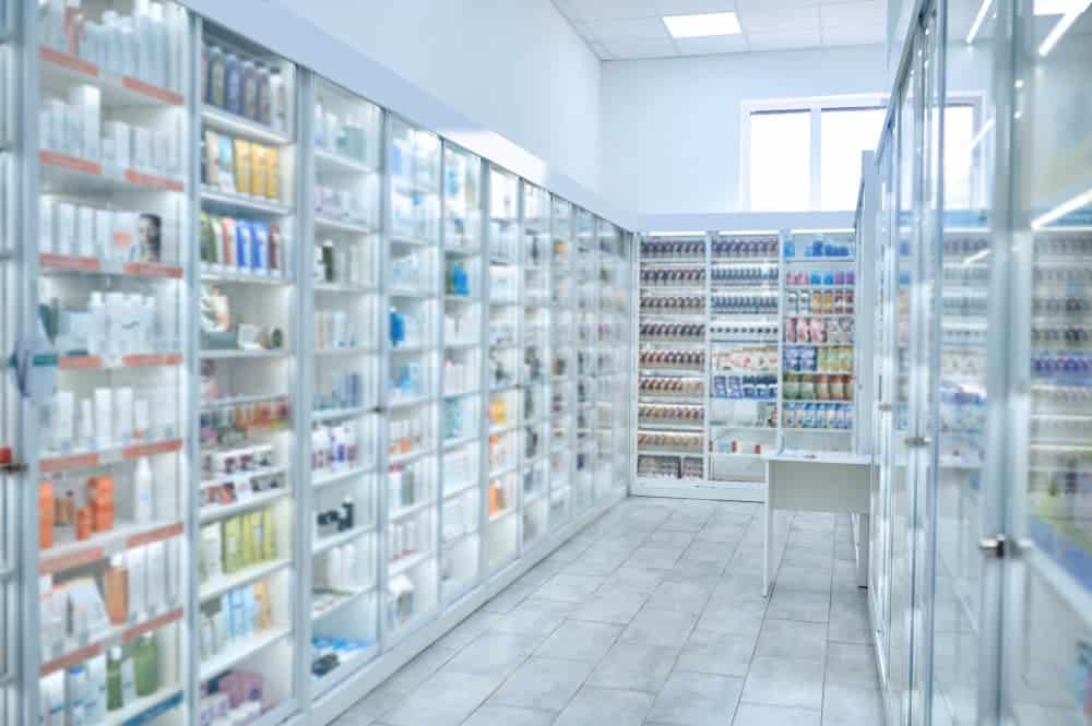 L'intérieur d'une pharmacie bien éclairée, avec des étagères garnies de divers médicaments, produits de santé et articles de toilette, et une petite table blanche dans le couloir.