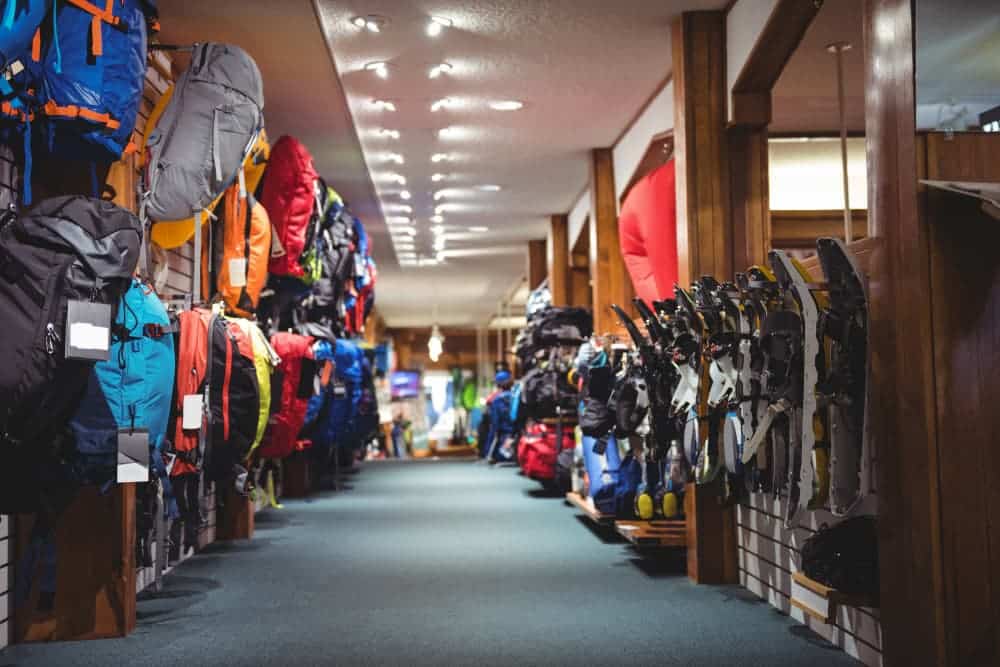 Une allée de magasin présentant des sacs à dos sur le mur de gauche et divers équipements de plein air, notamment des bâtons de ski et des casques, sur le mur de droite.