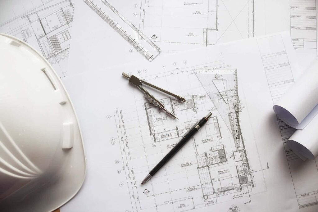 Plans d'architecte avec un stylo, un compas, une règle et un casque blanc sur un bureau ; des plans enroulés sont également visibles.