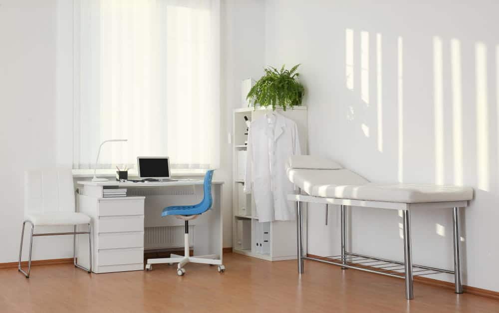 Un cabinet médical propre et moderne avec un bureau, des chaises, un ordinateur, une blouse blanche, une plante en pot et une table d'examen contre un mur blanc.