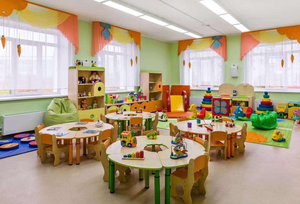 Salle de classe préscolaire avec de petites tables rondes, des chaises colorées, des étagères de jouets, des tapis de jeu et des décorations lumineuses. De grandes fenêtres laissent entrer la lumière naturelle.